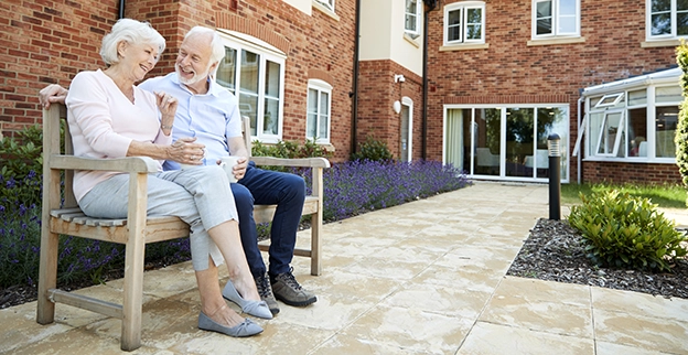 An older couple sitting on a bench outside of a senior living facility.